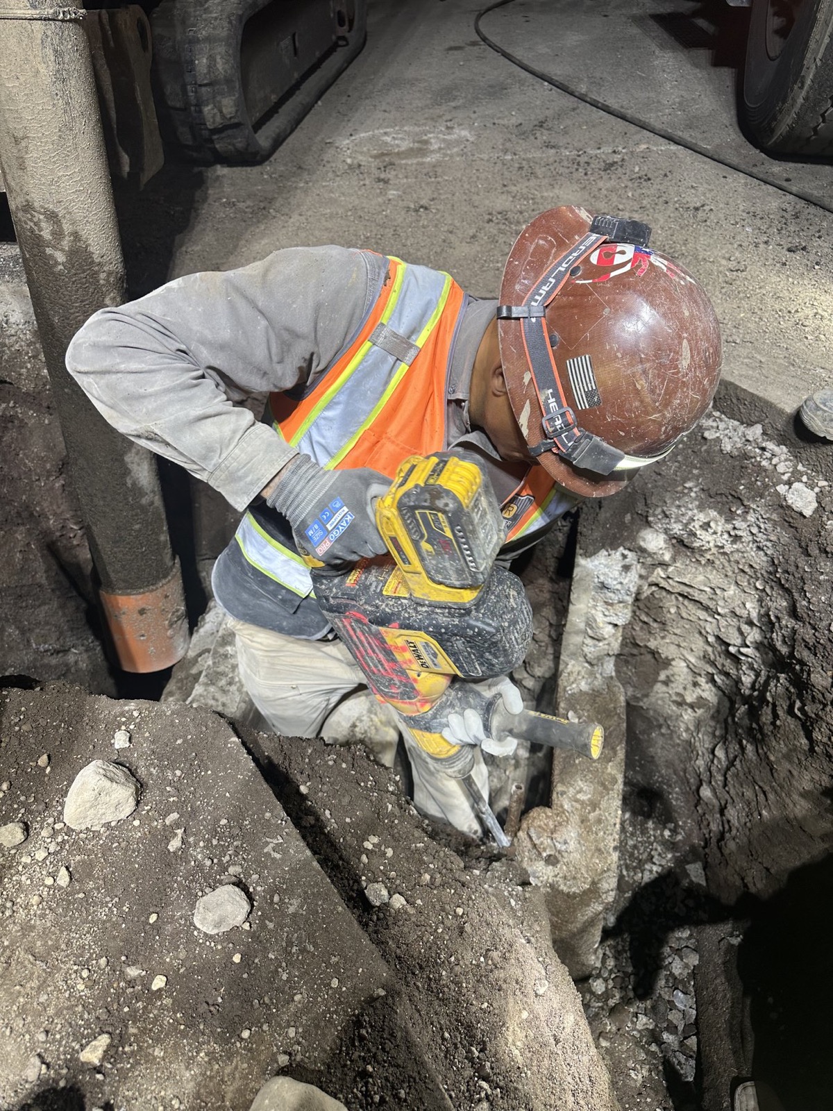 DeWalt 60V cordless SDS Max rotary hammer with 20V battery being used to break concrete around utility pipe in a deep trench, worker in hi-vis and hard hat